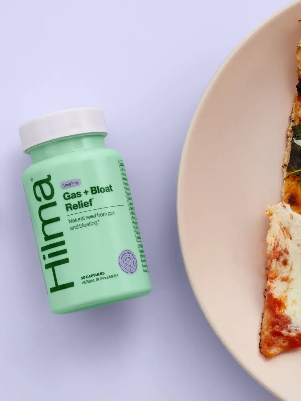 A bottle of Hilma Gas + Bloat Relief capsules is placed next to a slice of pizza on a plate.