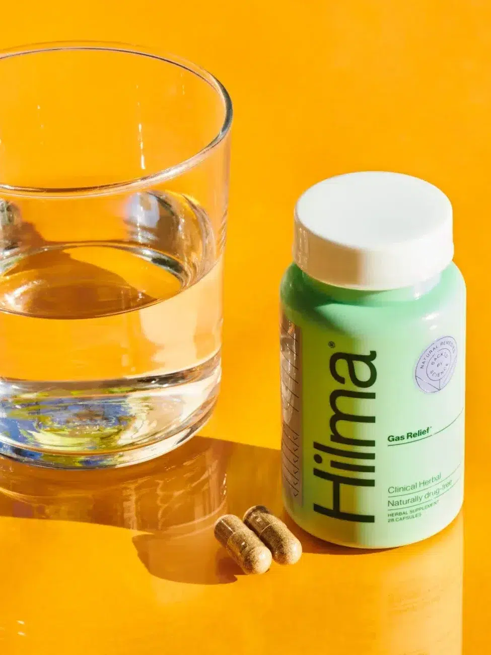 A glass of water next to a bottle of Hilma Gas Relief supplements and two capsules on a yellow surface.