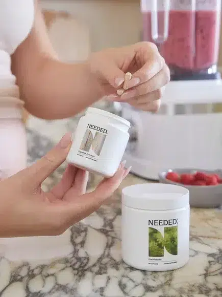 A person stands in a kitchen holding a pill with one hand and a white supplement jar labeled "Needed" with the other, with a smoothie blender and various fruits in the background.