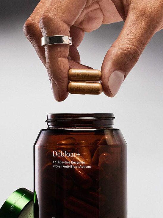 A hand holds two capsules above an open brown bottle labeled "Debloat+" with the subtitle "17 Digestive Enzymes Proven Anti-Bloat Actives." The bottle cap is off and placed on the side.