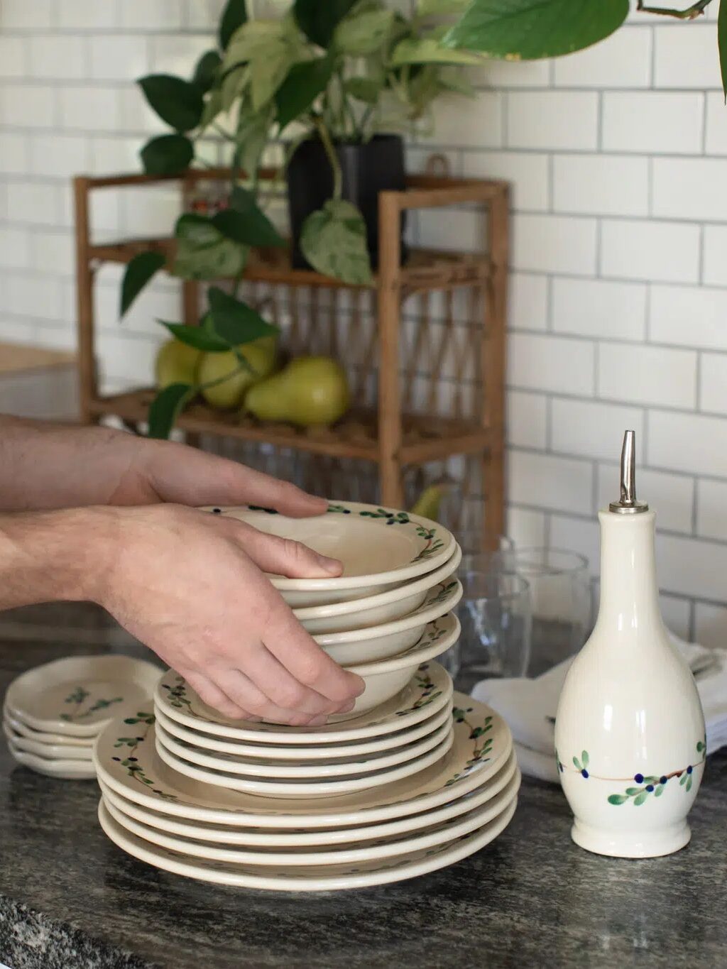 Person stacking a set of clean plates on a kitchen counter with plants and an olive oil dispenser nearby.