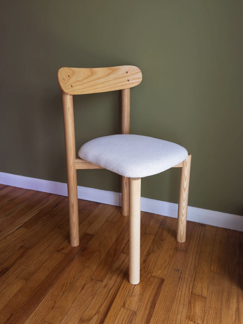 A wooden chair with a light-colored cushioned seat is placed on a hardwood floor against a green wall.