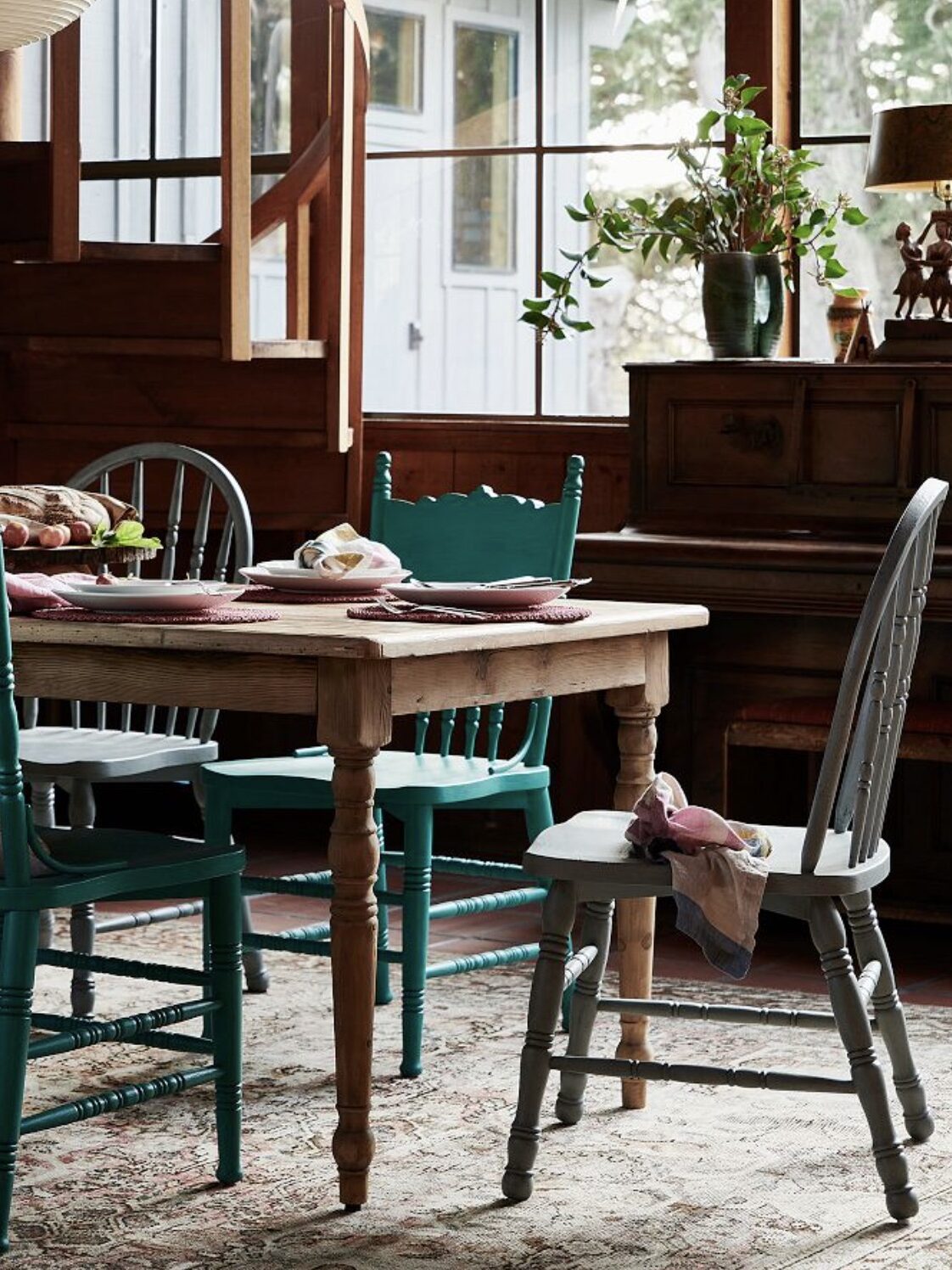 A rustic dining room with a wooden table, assorted chairs, tableware, and a plant on a sideboard. Sunlight filters through windows, illuminating the cozy space decorated with homey accents.