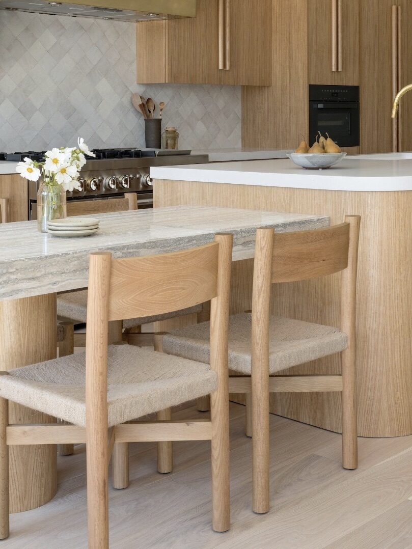 Modern kitchen with light wood cabinets, a large island with a white countertop, and matching wooden chairs. A vase with white flowers and a bowl with fruits are on the island. A range hood is visible.