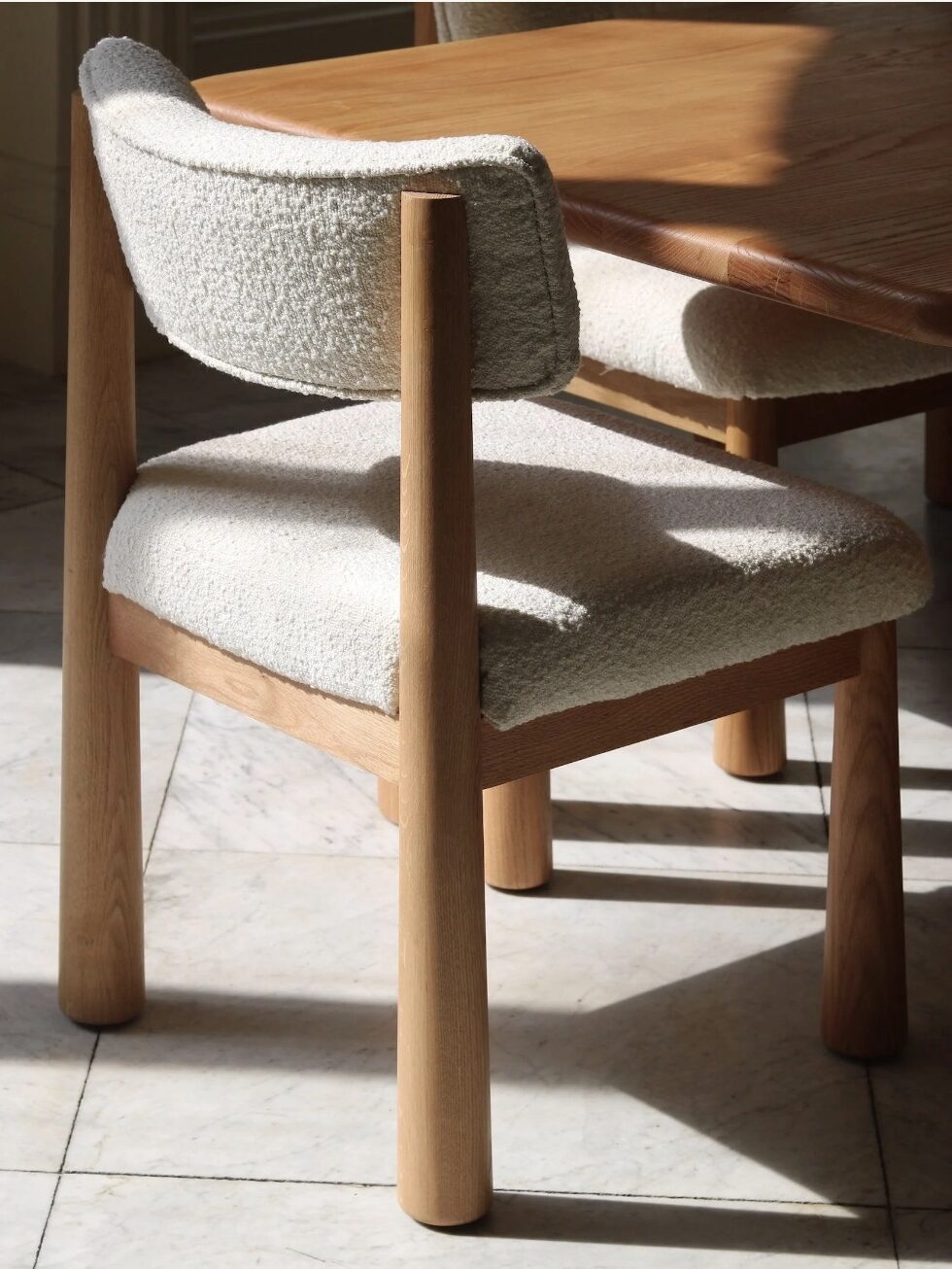 A wooden chair with a light-colored cushioned seat and backrest is positioned next to a wooden table on a tiled floor.