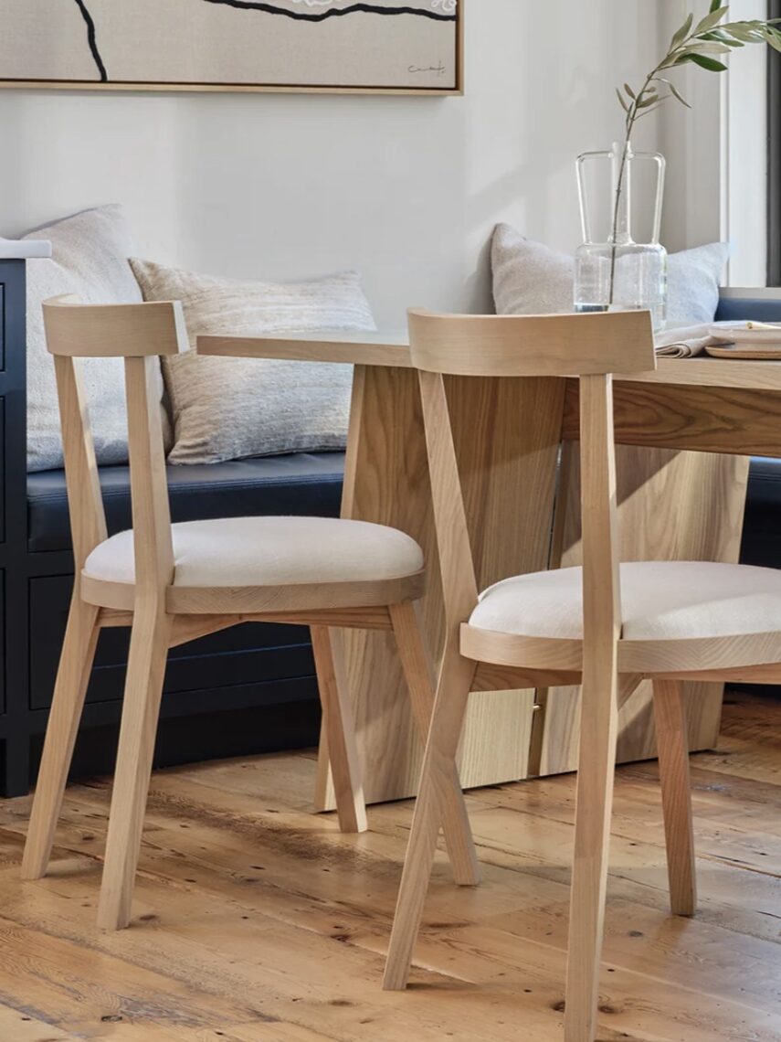 A modern wooden dining table set with two chairs, a carafe, and glassware in a bright room with a window and cushioned seating area.