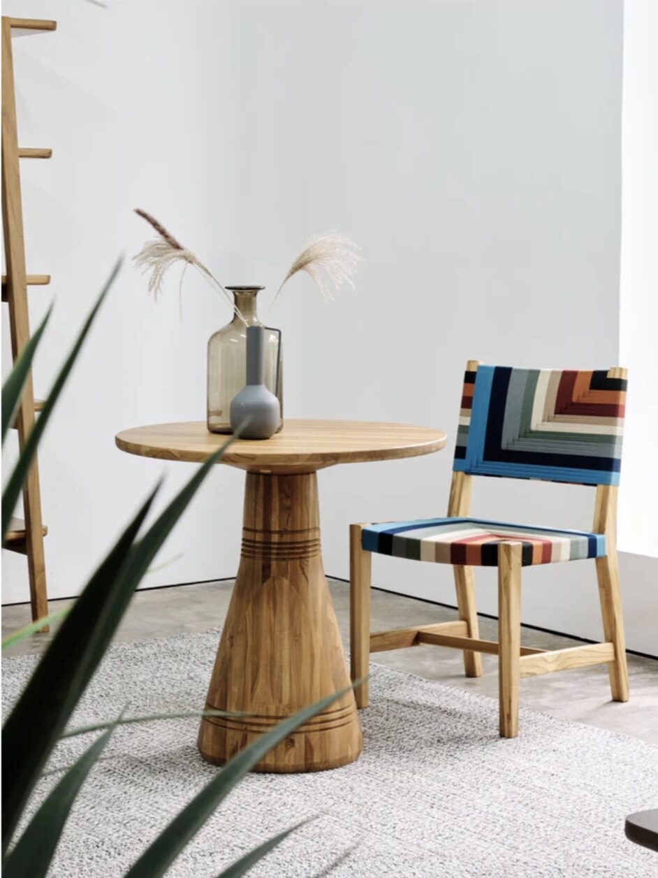 A small round wooden table with a modern two-tone vase and a decorative chair with a colorful, striped pattern in a minimalist room.