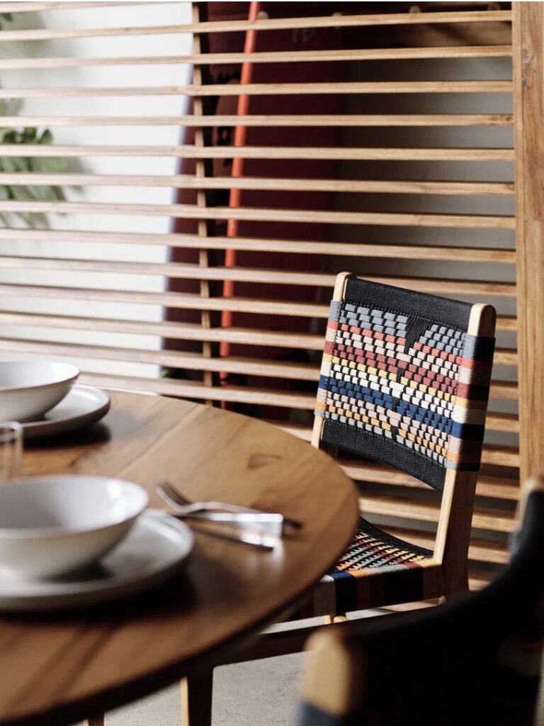 A dining table set with plates, bowls, and cutlery, beside a wooden slatted divider. Behind it, a colorful woven chair is visible.