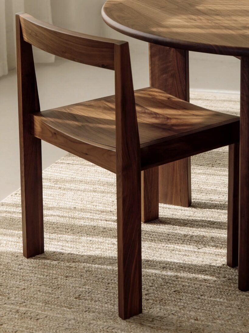 A wooden chair with a simple design is positioned next to a round wooden table on a beige rug, in a well-lit room.