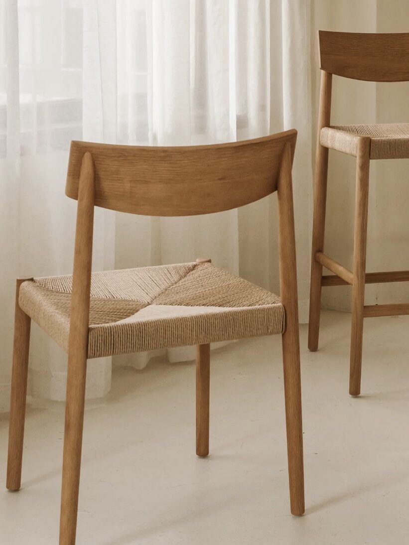 Two wooden chairs with woven seats are placed on a light-colored floor in front of sheer white curtains.