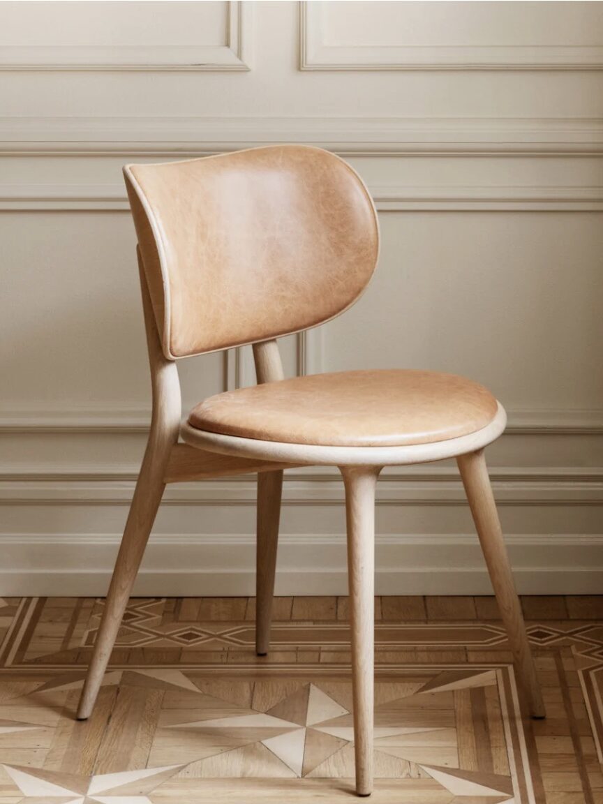 A wooden chair with a round seat and curved backrest, upholstered in light brown leather, placed on a wooden patterned floor against a cream-colored paneled wall.