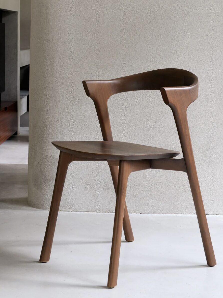 A modern wooden chair is placed on a light-colored floor against a plain wall. The chair features clean lines and a curved backrest.