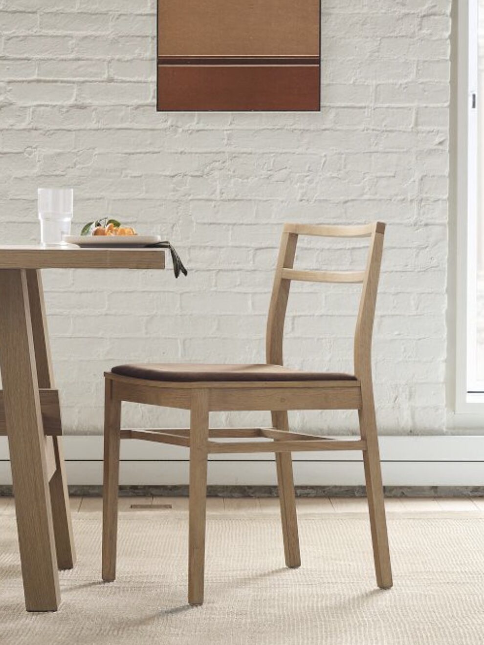 A wooden chair with a dark seat cushion placed at a dining table set against a white brick wall. A partial view of a meal, glass, and artwork can also be seen.