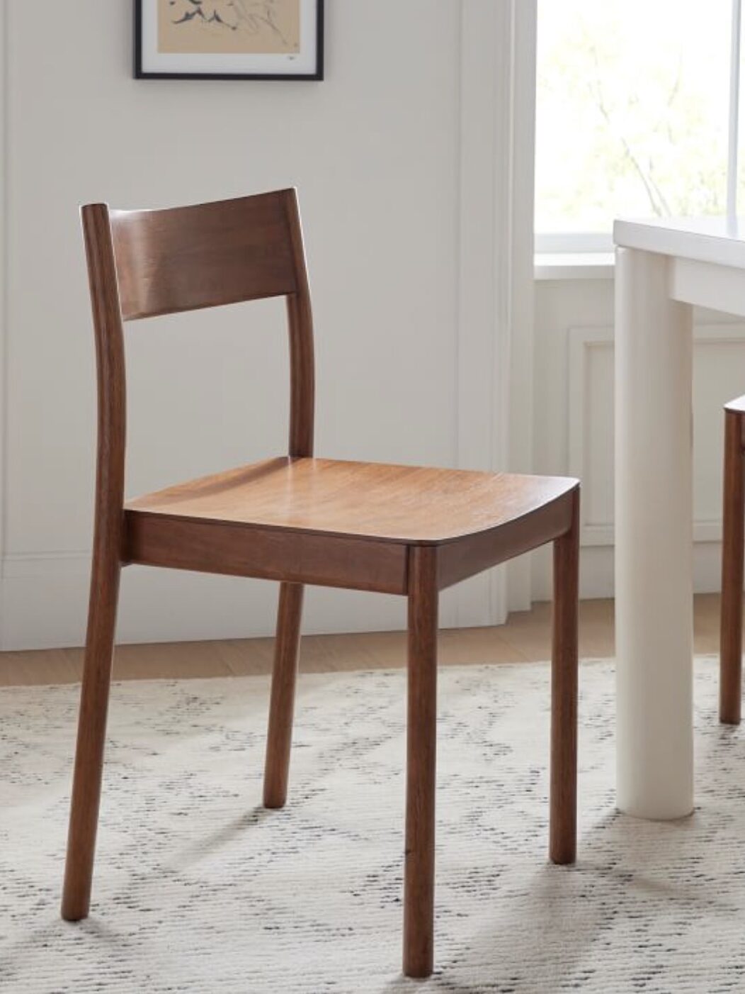 A wooden chair with a straight backrest is placed on a light-colored rug next to a white table in a bright, sunlit room.