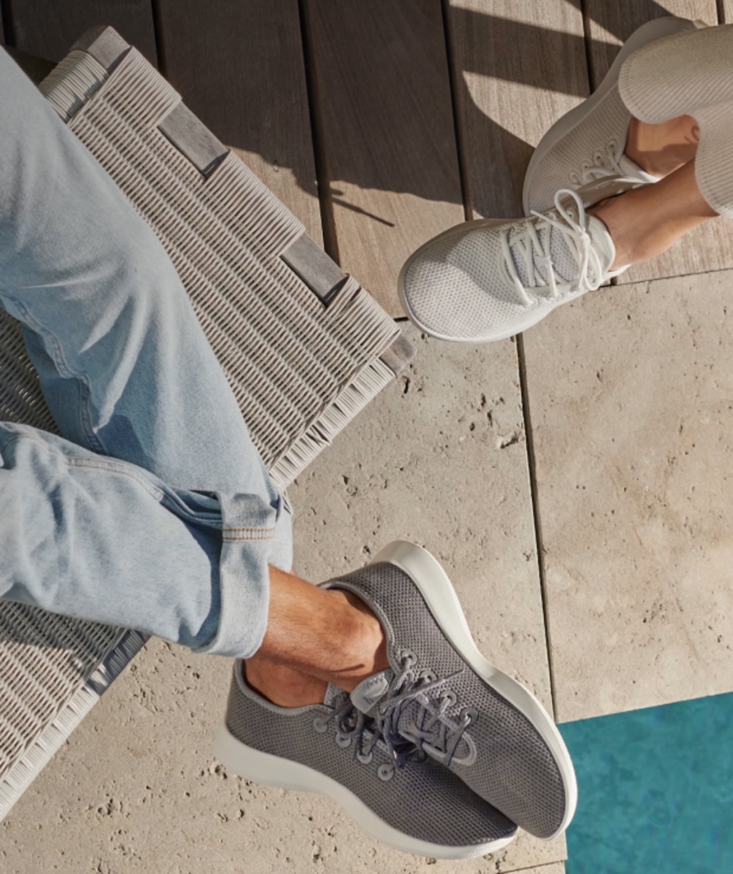 Two people wearing casual sneakers sit on a deck near a pool, with one wearing light gray shoes and jeans and the other in light-colored shoes and pants.