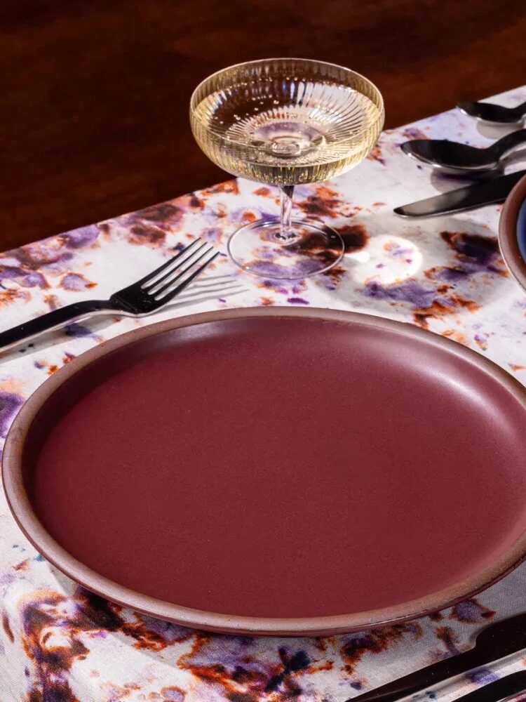 A table setting with a glass of sparkling wine, silverware, and layered ceramic dishes in burgundy and blue hues on a floral tablecloth.