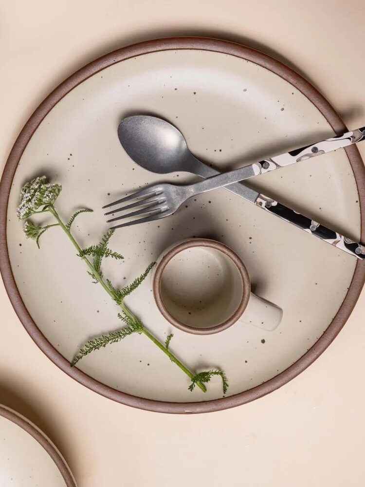 A ceramic plate with a silver spoon and fork, a small cup, and a sprig of greenery. The utensils have black and white handles. All items reside on a beige surface.