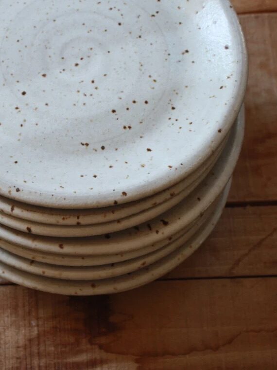 A stack of eight white ceramic plates with brown speckles, neatly arranged on a wooden surface.