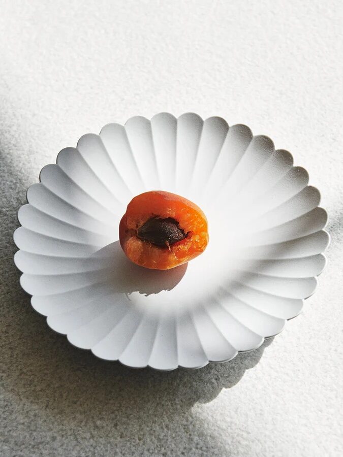 Half of an apricot with a stone on a white, scalloped plate, under soft sunlight.