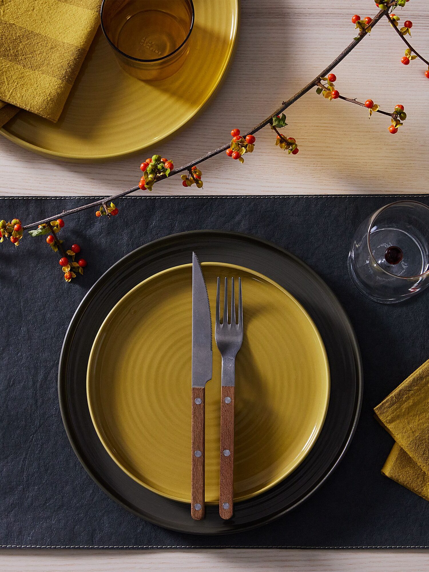 A yellow plate with a fork and knife is placed on a dark placemat. A glass with red liquid and a napkin are beside it. Decorative branches with berries are above the setting.