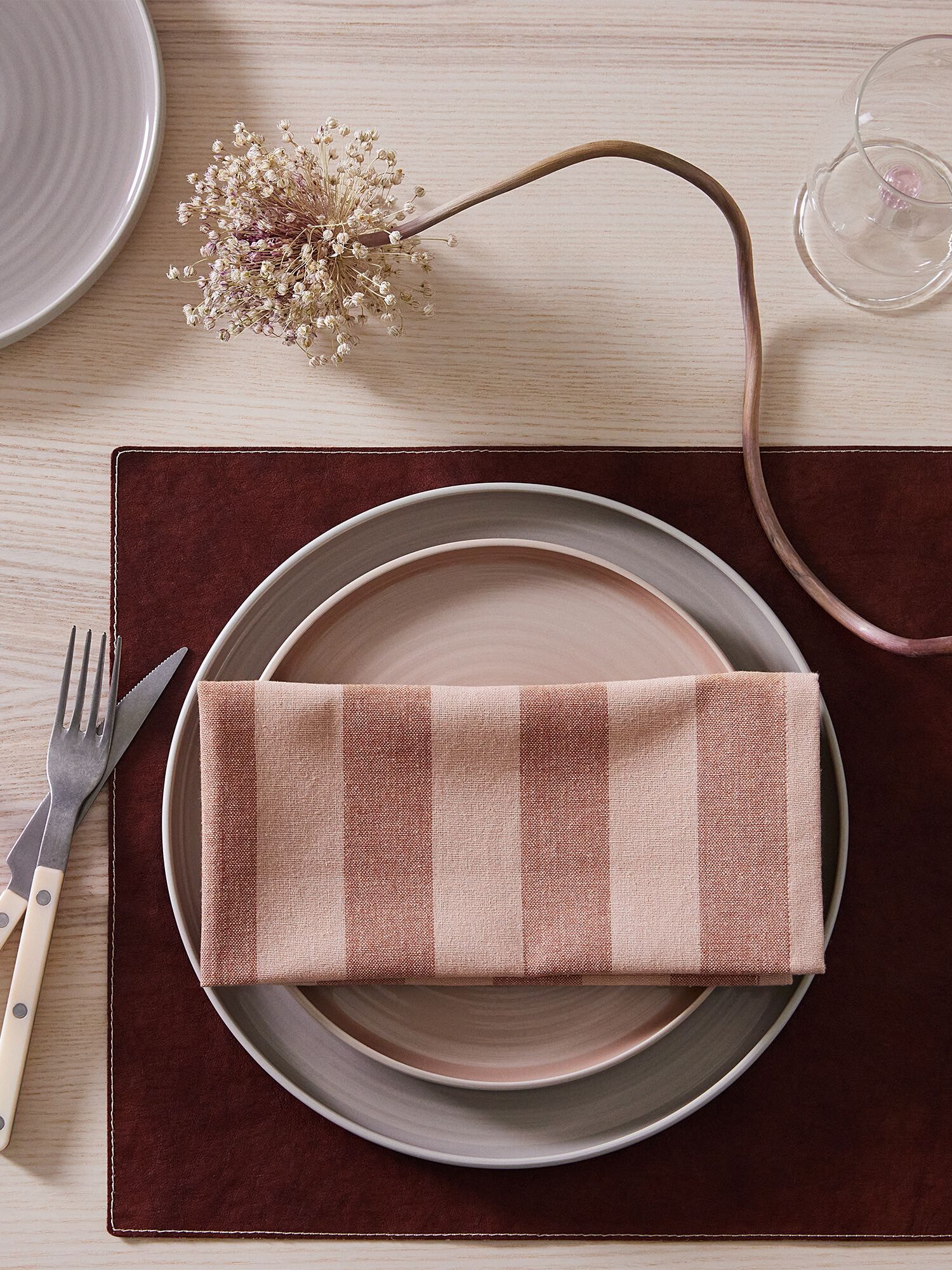 A neatly arranged table setting with a pink striped napkin on a plate, alongside a fork and knife, a glass, a pink cup, and a small bouquet on a light wood surface.