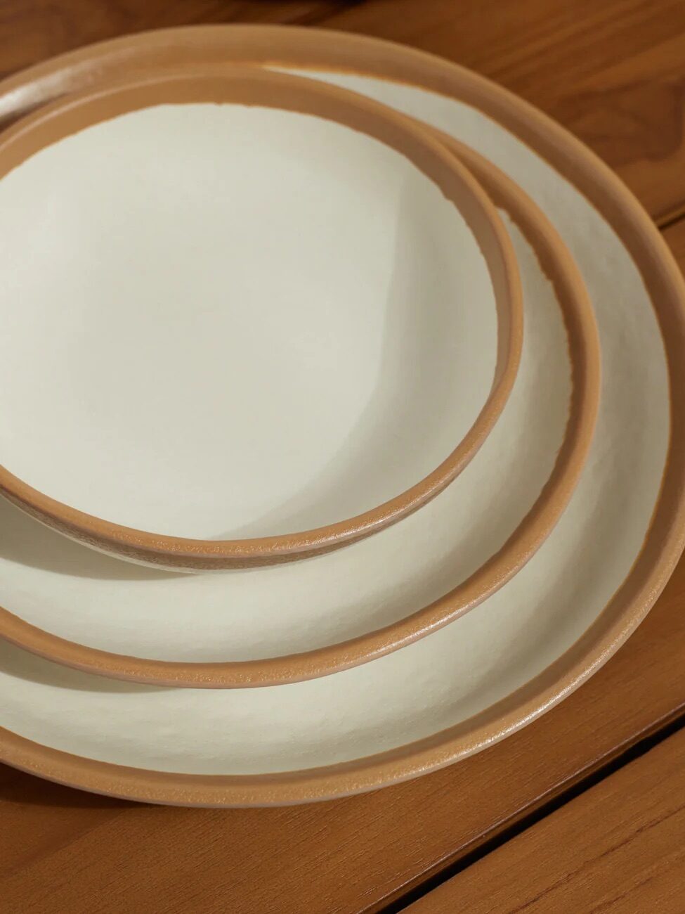 Three nested ceramic plates, each with a white interior and light brown rim, are stacked on a wooden surface.