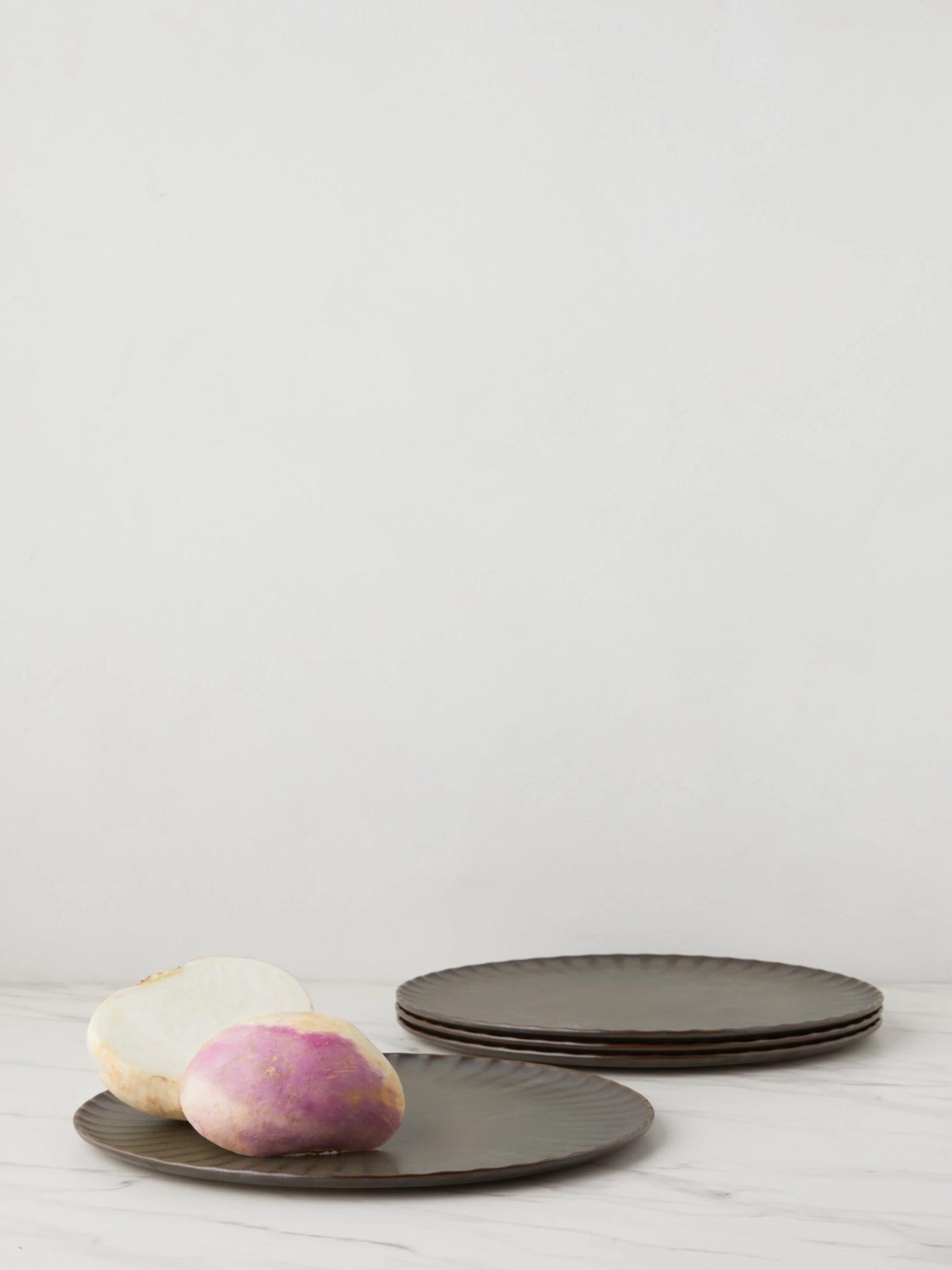 A sliced turnip and a whole turnip are placed beside a stack of dark plates on a light-colored surface against a plain background.