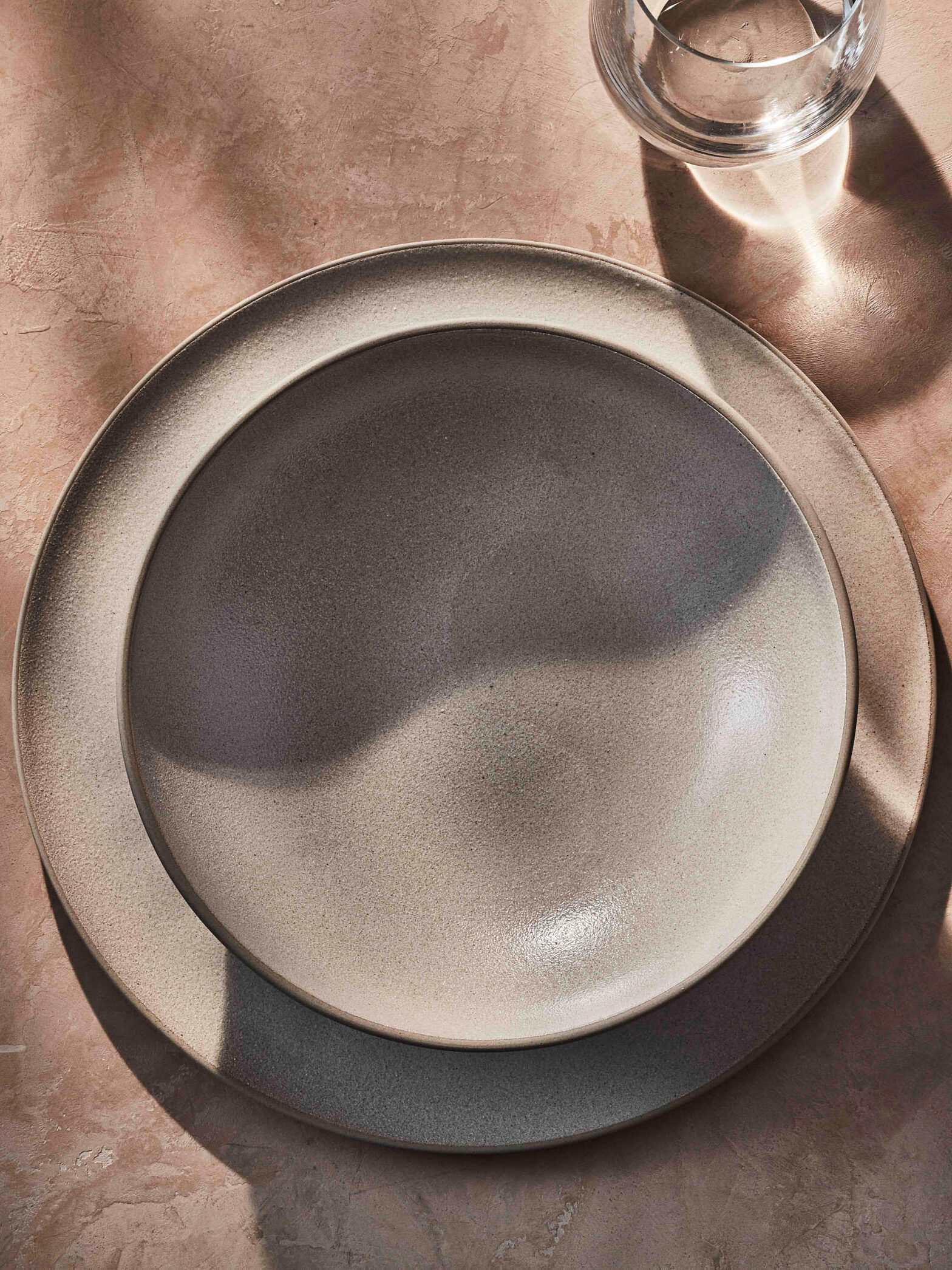 A minimalist table setting featuring a grey plate on another grey plate, a fork to the left, a glass and spoon to the right, and a knife above the plate, all on a textured surface.