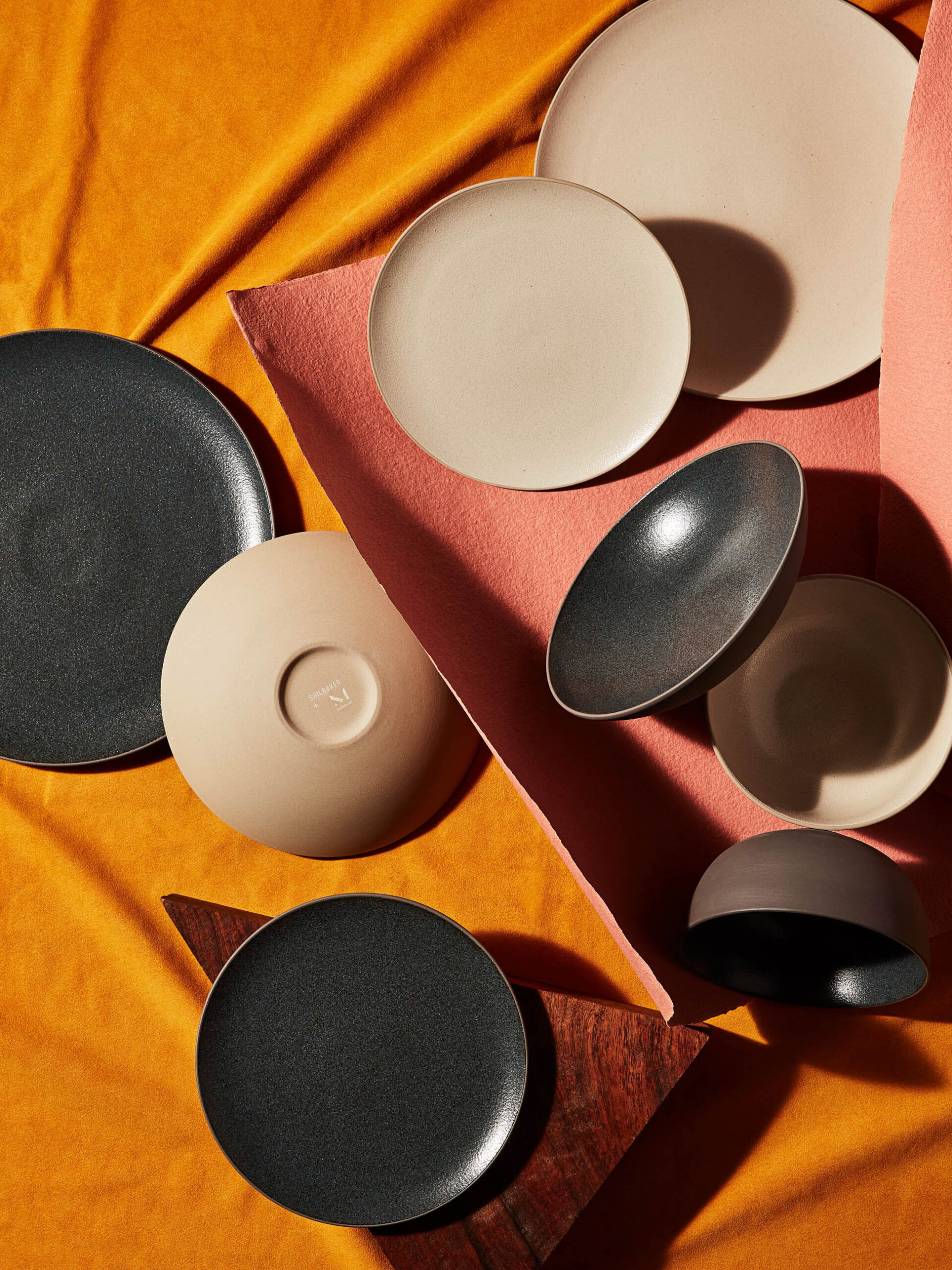 A variety of black, beige, and gray ceramic bowls and plates are artistically arranged on a yellow and pink fabric background.