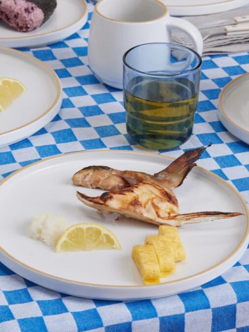 A grilled fish head, pieces of omelette, lemon wedge, and white garnish on a white plate with other dishes on a blue and white checkered tablecloth.