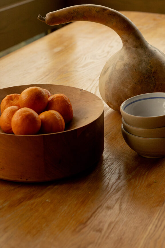 A wooden table with a bowl of oranges, a stack of bowls, and a sculpture on it.