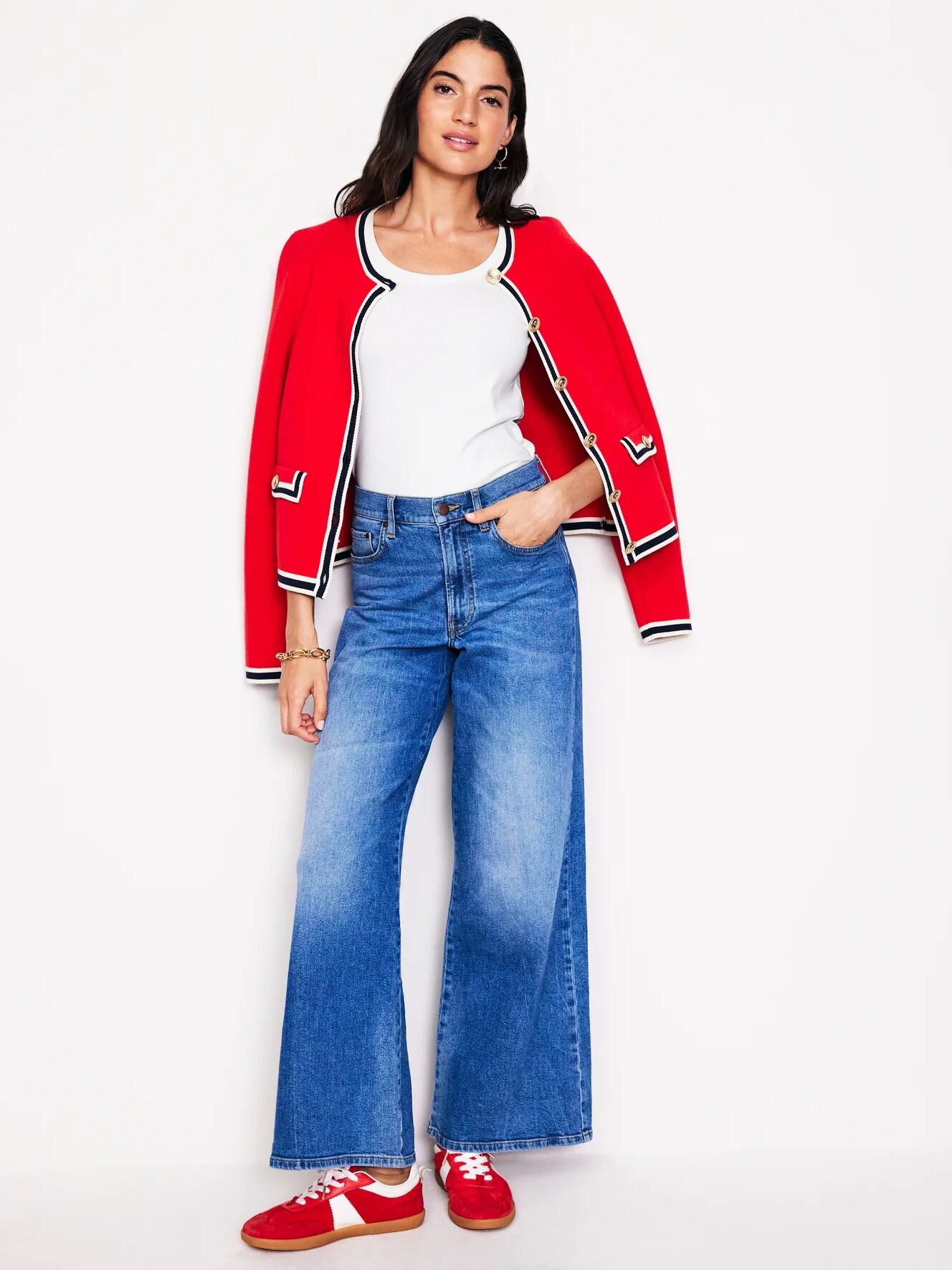 Woman in wide-leg jeans, white top, and red jacket, poses with one hand in her pocket. She is wearing red sneakers and standing against a plain white background.