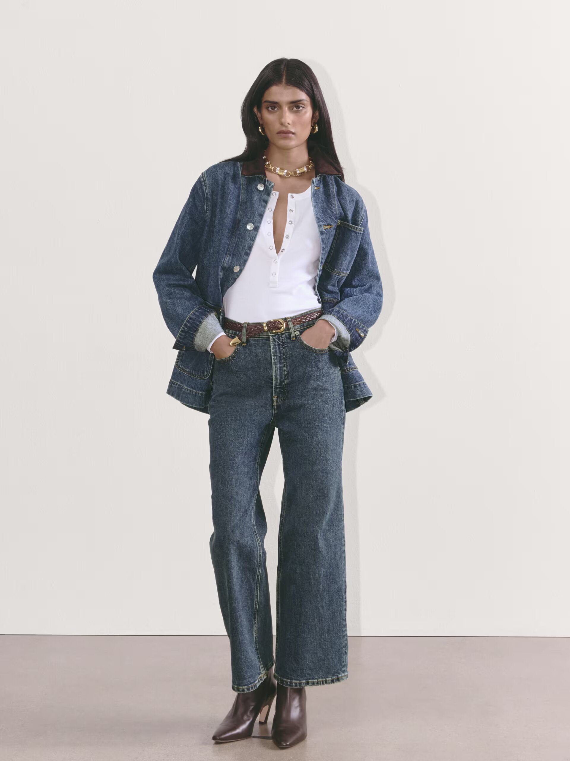 A person stands against a plain background wearing a white top, denim jacket, jeans, a belt, brown boots, and jewelry. Hands are in the pockets of the jeans.