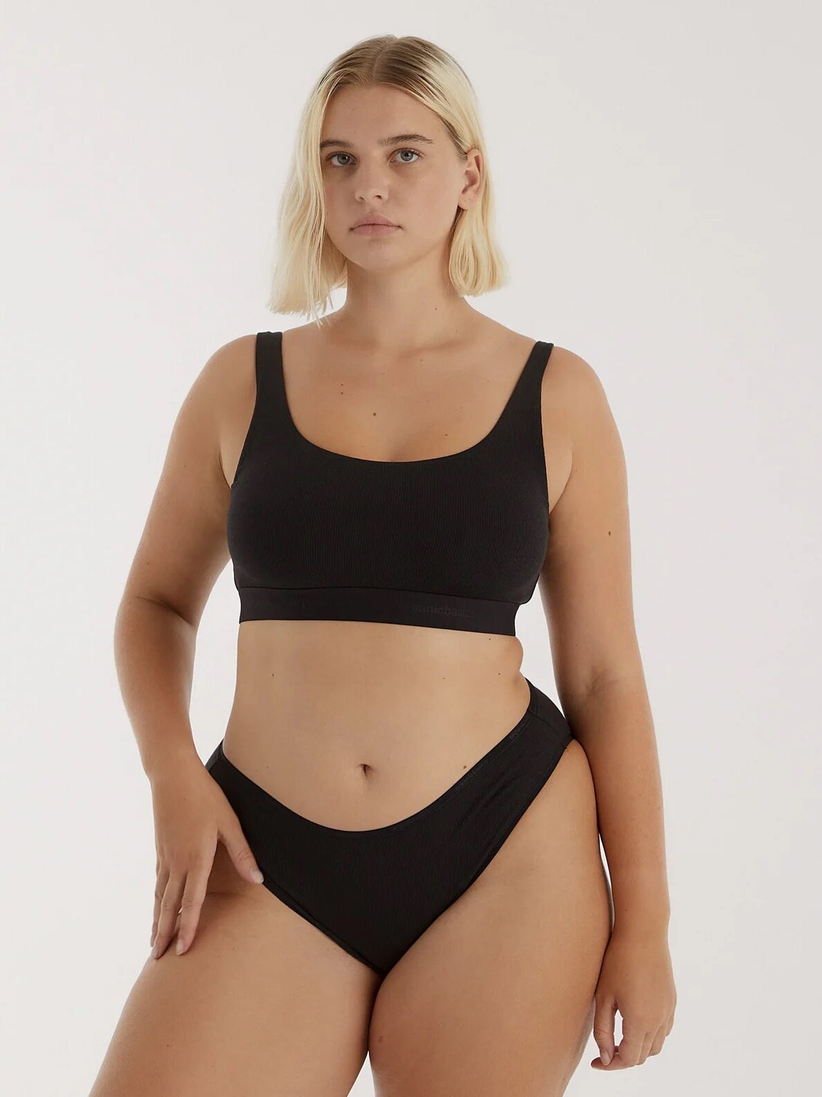 A person stands in a plain setting wearing a matching black bikini, with a neutral facial expression and hands resting on the body.