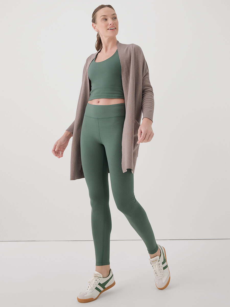 A woman wearing a green sports bra and leggings set with a taupe cardigan and white sneakers stands against a plain white background.