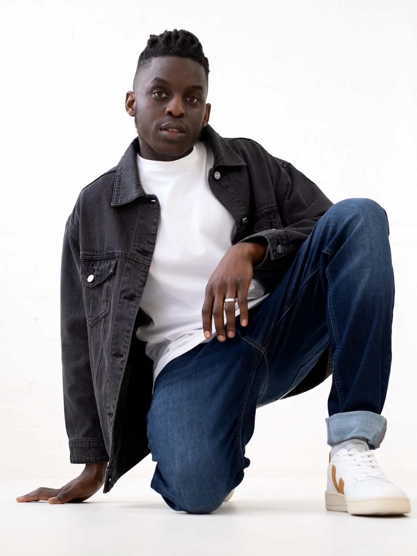 Person kneeling on the ground, wearing a white T-shirt, black denim jacket, blue jeans, and white sneakers, with a white background.