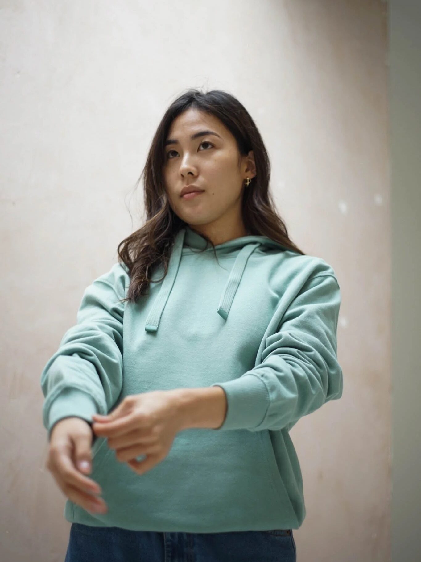 Woman in a light green hoodie stands indoors, adjusting her sleeves with a neutral expression.