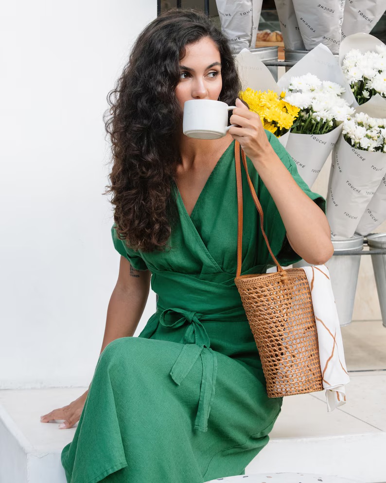 A woman in a green dress sips from a white mug while sitting with a woven bag beside her.