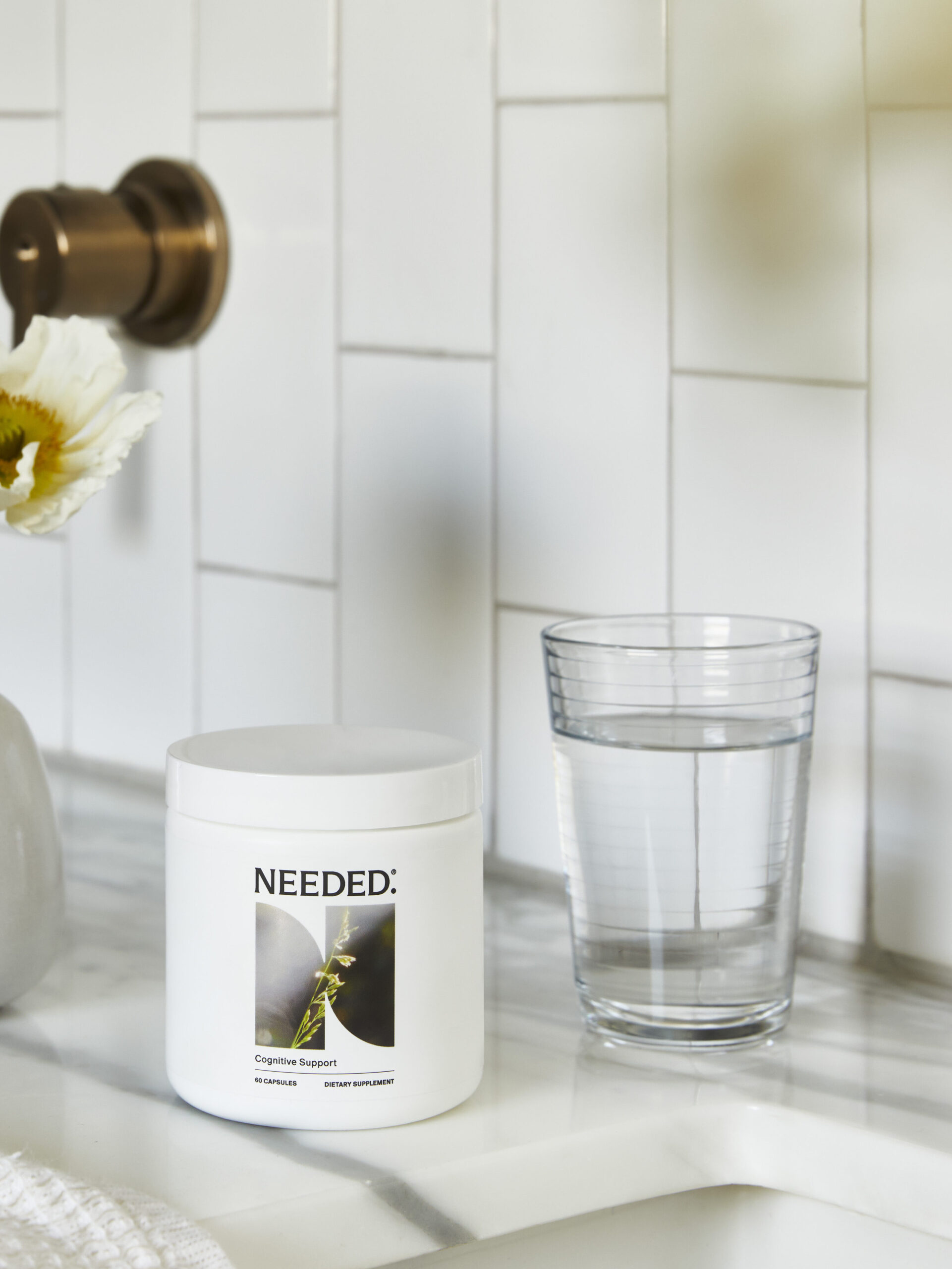 A jar labeled "Needed," a vase with white flowers, and a glass of water are arranged on a white tiled bathroom countertop next to a bronze faucet.