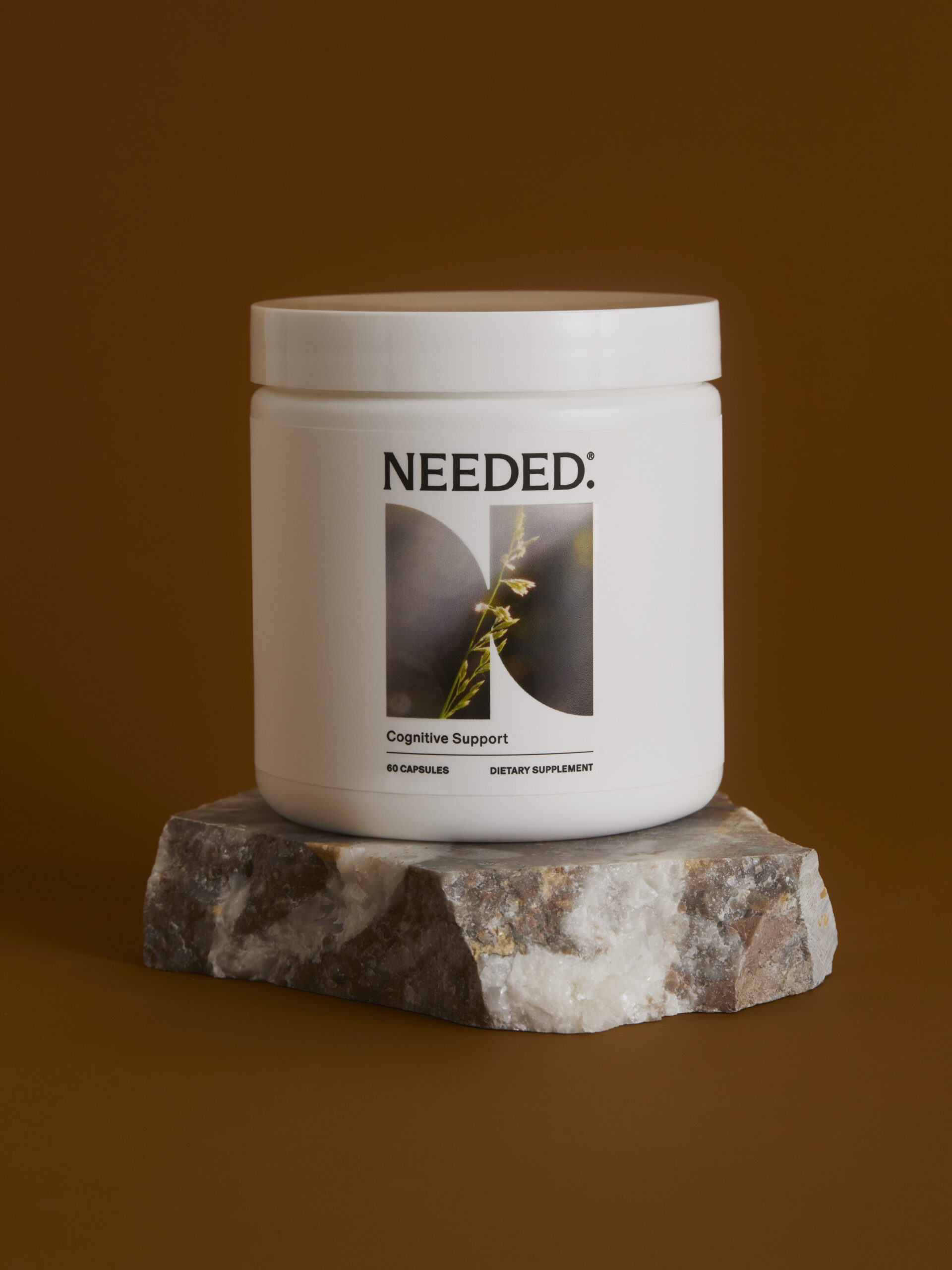 A white jar labeled "NEEDED: Cognitive Support" with 60 capsules, dietary supplement, is placed on a stone pedestal against a brown background.