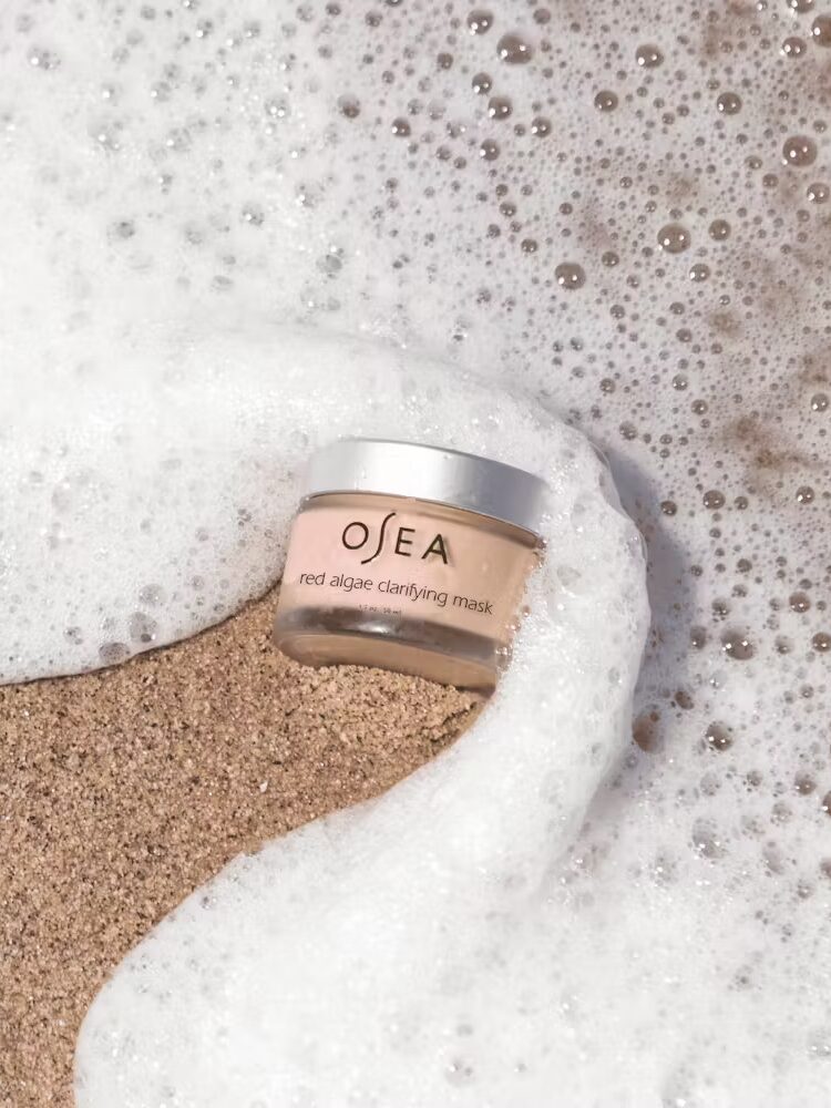 A jar of Osea red algae clarifying mask is partially covered by sea foam on a sandy beach.