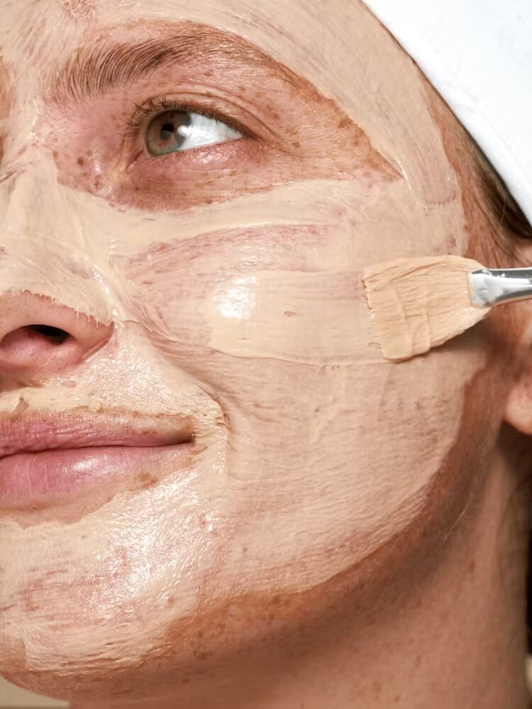 Close-up of a person with a clay facial mask being applied to their face using a brush. They are wearing a white headband and have a partial smile.