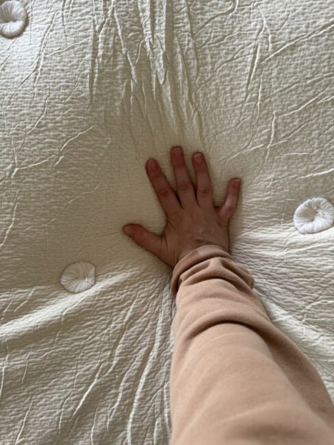 A hand is pressing down on a textured latex mattress, creating an indentation. The person is wearing a beige long-sleeve shirt.