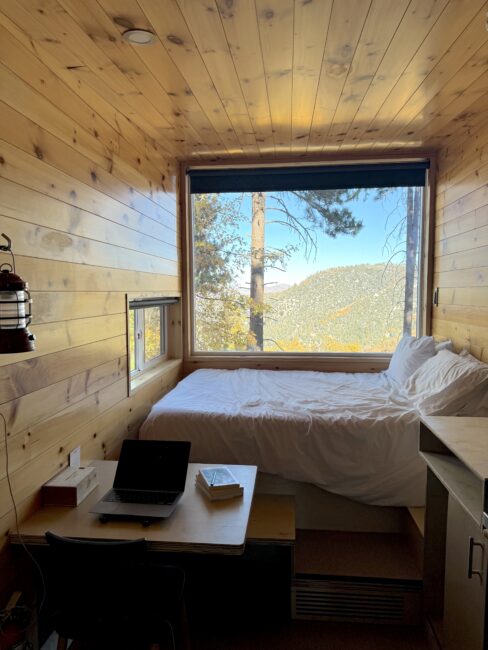 A small wooden cabin interior with a large window facing mountains and trees. The space includes a bed, a desk with a laptop and book, and a lantern on the wall.