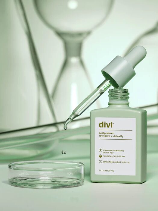 A dropper bottle of Divi scalp serum with a pipette and a small droplet of the serum, surrounded by lab glassware, on a green background. The bottle label mentions scalp revitalization and detox.