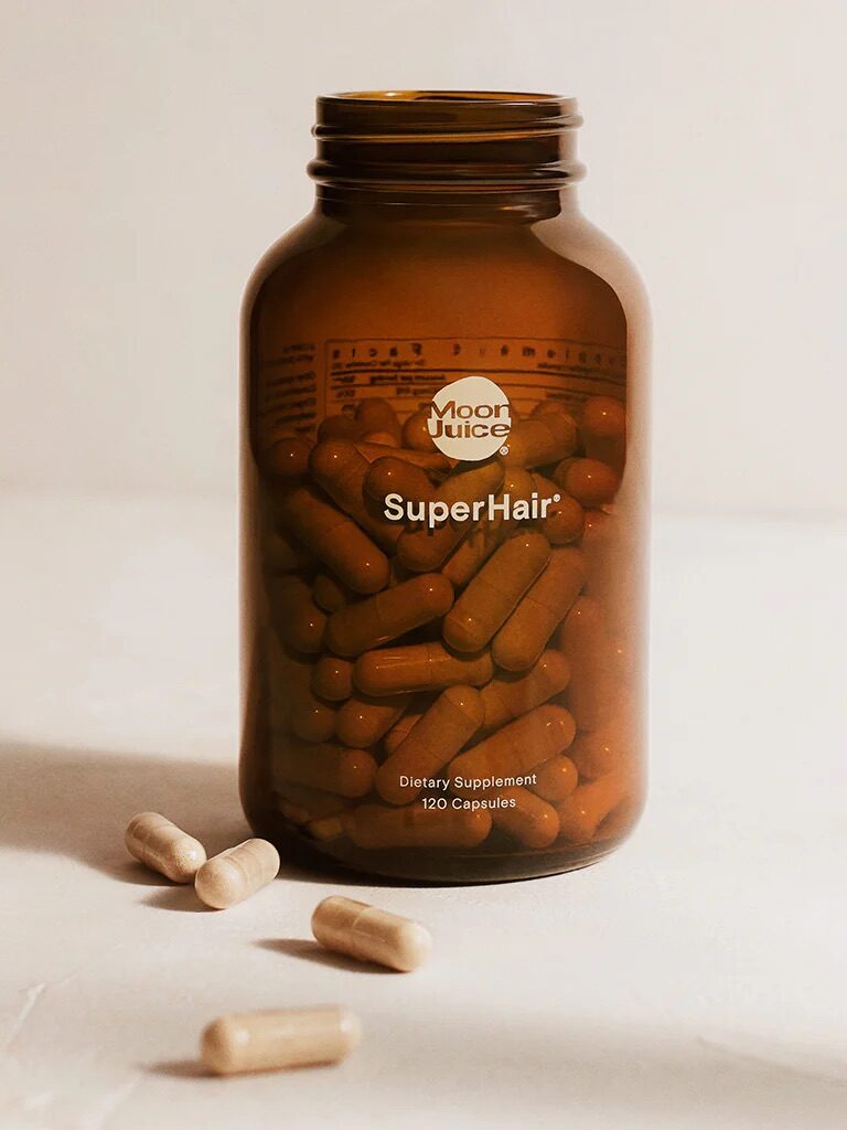 A brown plastic bottle labeled "Moon Juice SuperHair Dietary Supplement" with 120 capsules inside, and a few capsules scattered on the surface around it.