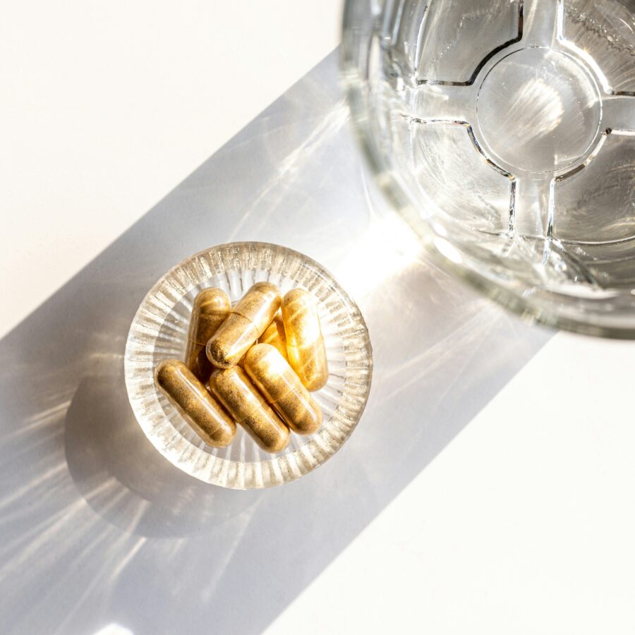 A small glass dish containing several gold capsules sits next to a glass of water on a white surface.