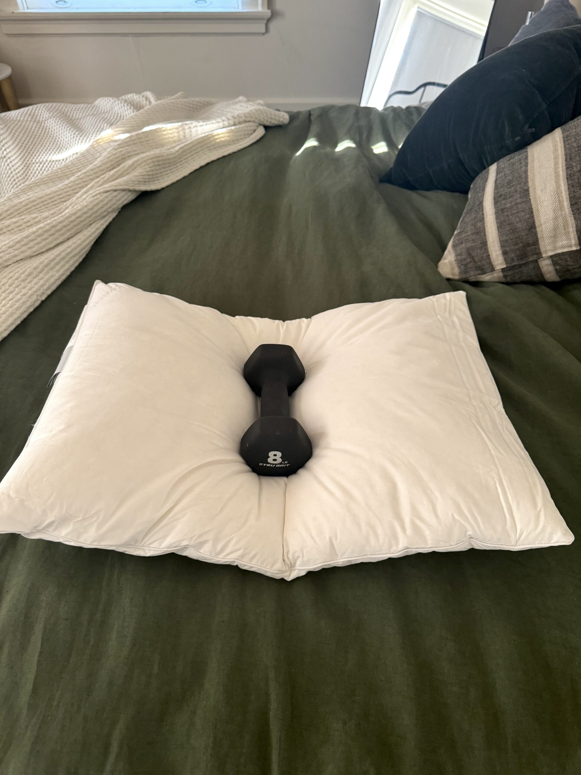 A black dumbbell resting on a white pillow on a green bedspread, with additional pillows and blankets in the background.