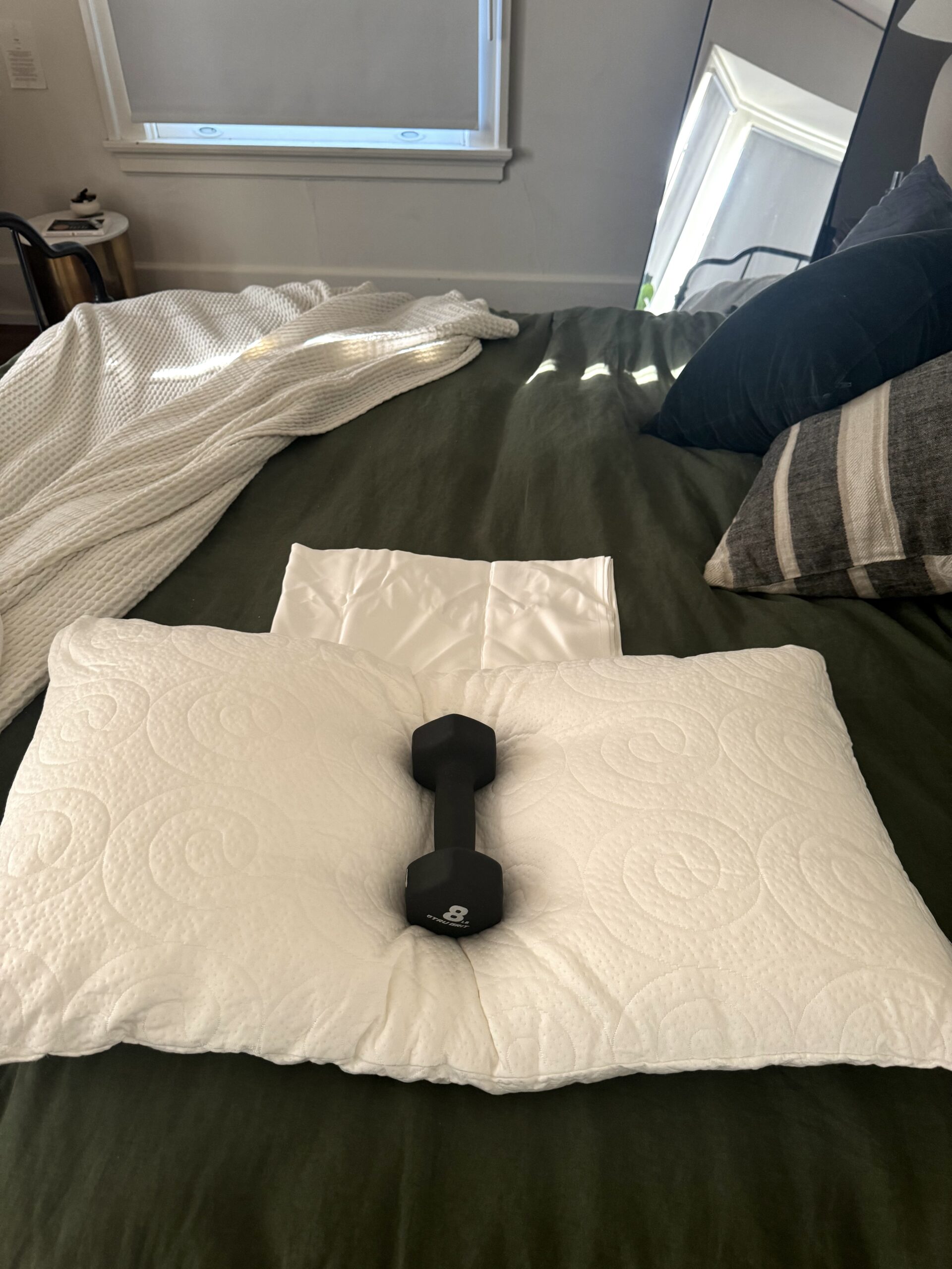 A white pillow and a black dumbbell are placed on a neatly made bed with a green blanket. A folded white blanket and a black-and-white striped pillow are also on the bed.