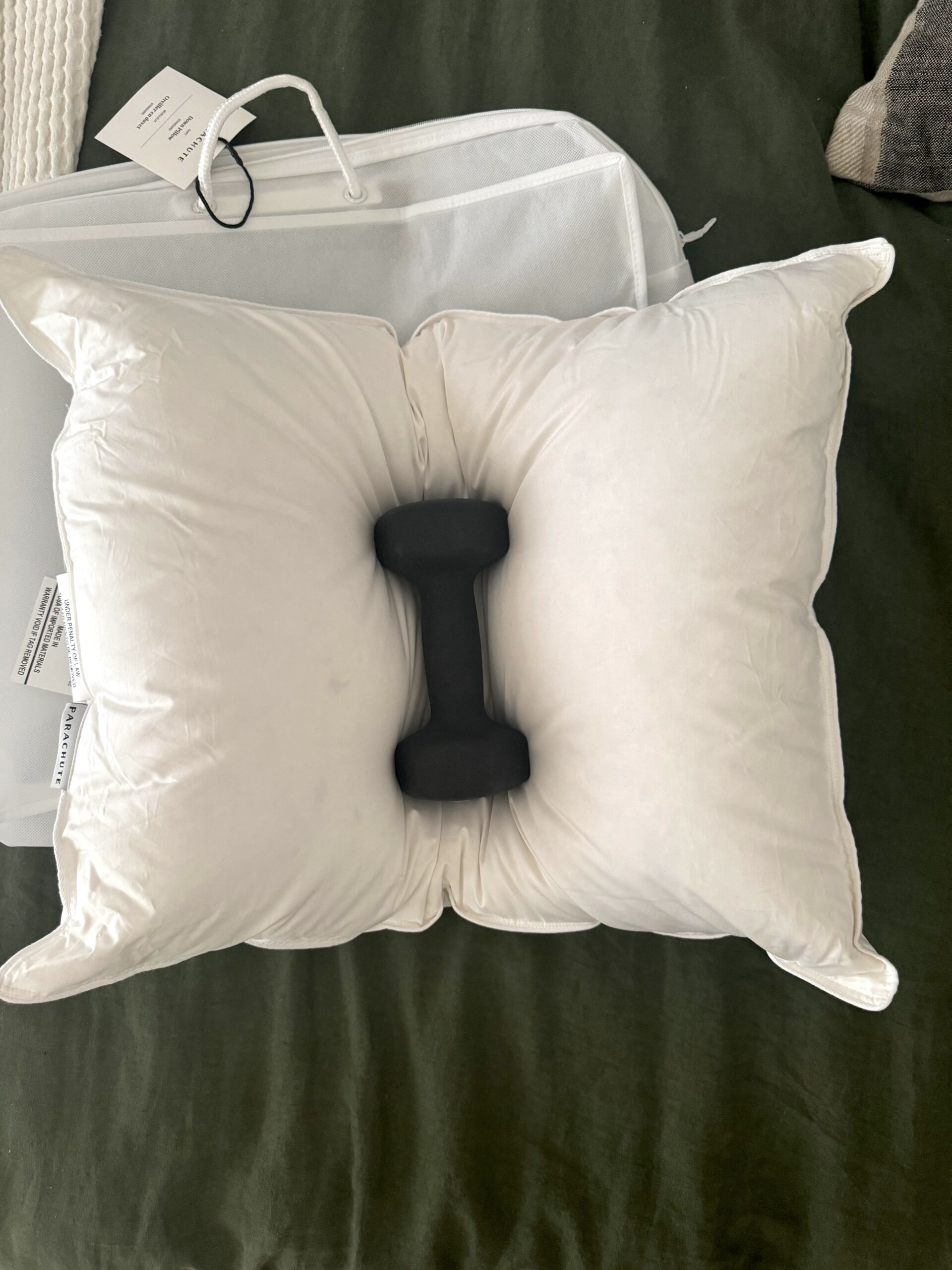 A black dumbbell resting in the middle of a white pillow placed on a dark green surface, with a mesh laundry bag and a white tag nearby.